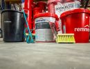 Rote Eimer mit &bdquo;Remmers BIT 1K&ldquo; und Reinigungswerkzeug stehen auf Boden, zeigen Bauchemie im Lager.